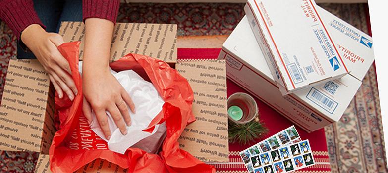 一个人正在包装一个假日包裹，桌上放着优先邮箱和假日邮票.